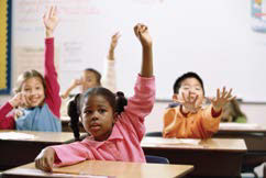 Photo of children at desks in a classroom with hands raised